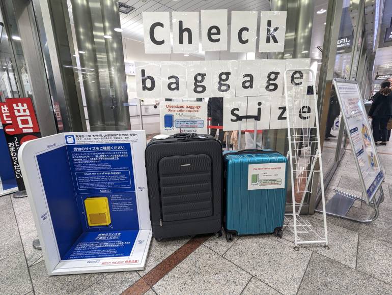 A black suitcase and a blue suitcase are next to a luggage sizer. On the window behind them a sign reads 'check baggage size'