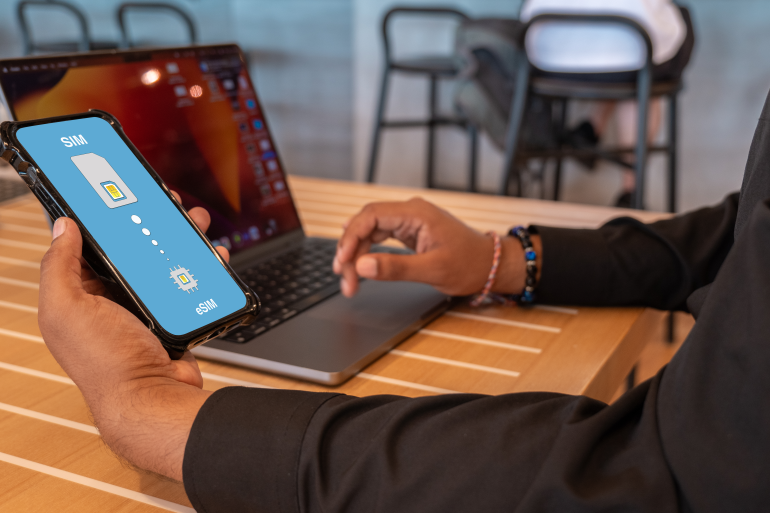 A man in business attire works at his laptop. He is holding a phone that has an eSIM installed.